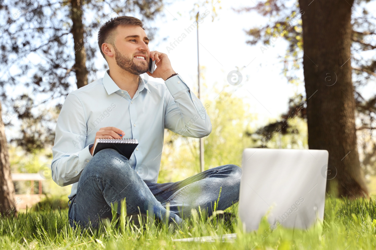 Image of Happy young man with laptop talking on phone in park 