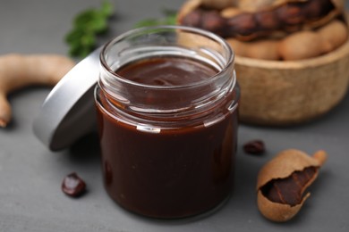 Photo of Jar of tasty tamarind sauce and fresh pods on grey wooden table, closeup
