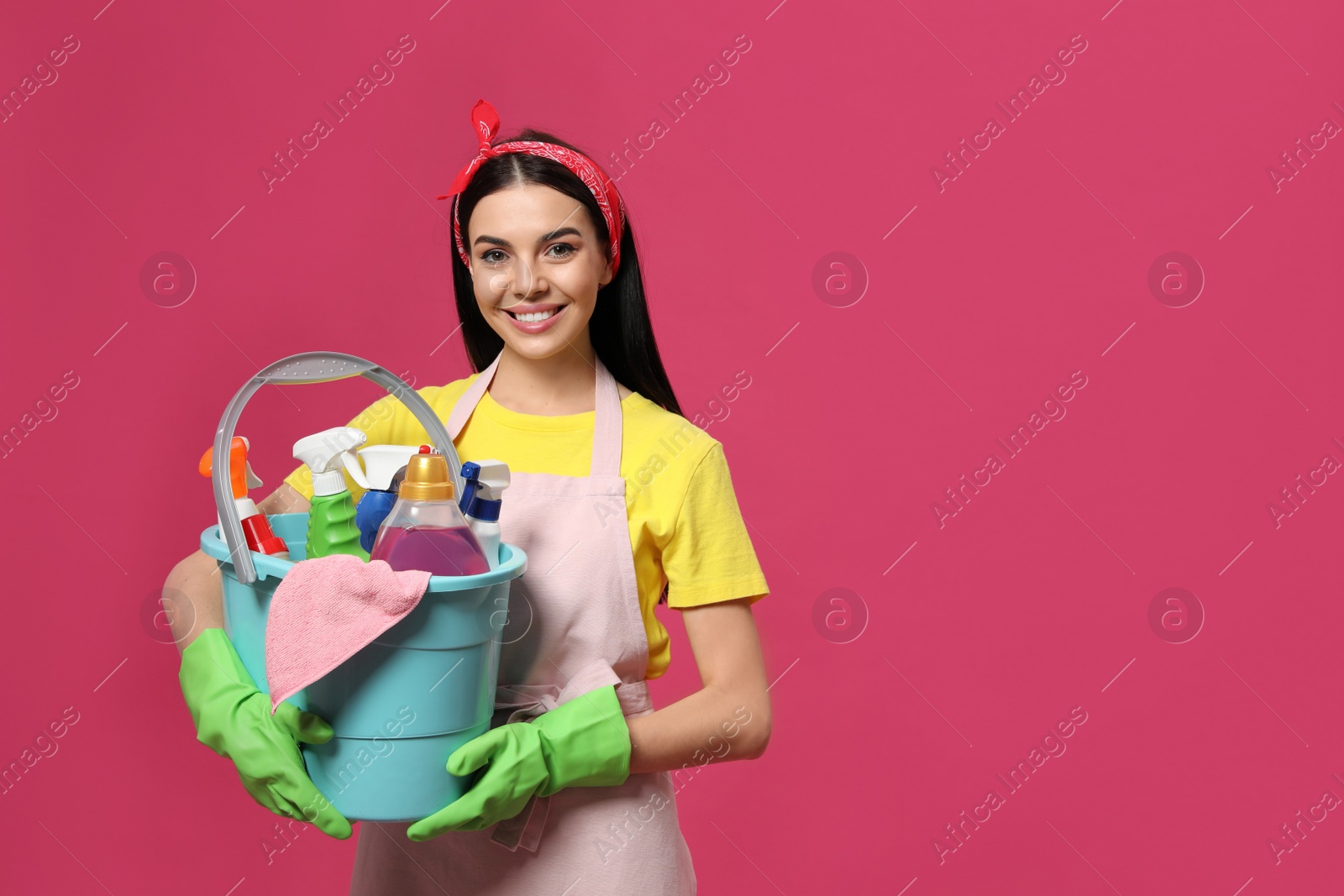 Photo of Young housewife holding bucket with cleaning supplies on pink background, space for text