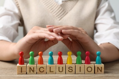 Photo of Woman protecting colorful pawns and wooden cubes with word Inclusion at table, closeup