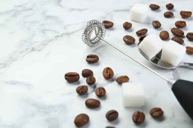 Photo of Black milk frother wand, sugar cubes and coffee beans on white marble table, closeup. Space for text