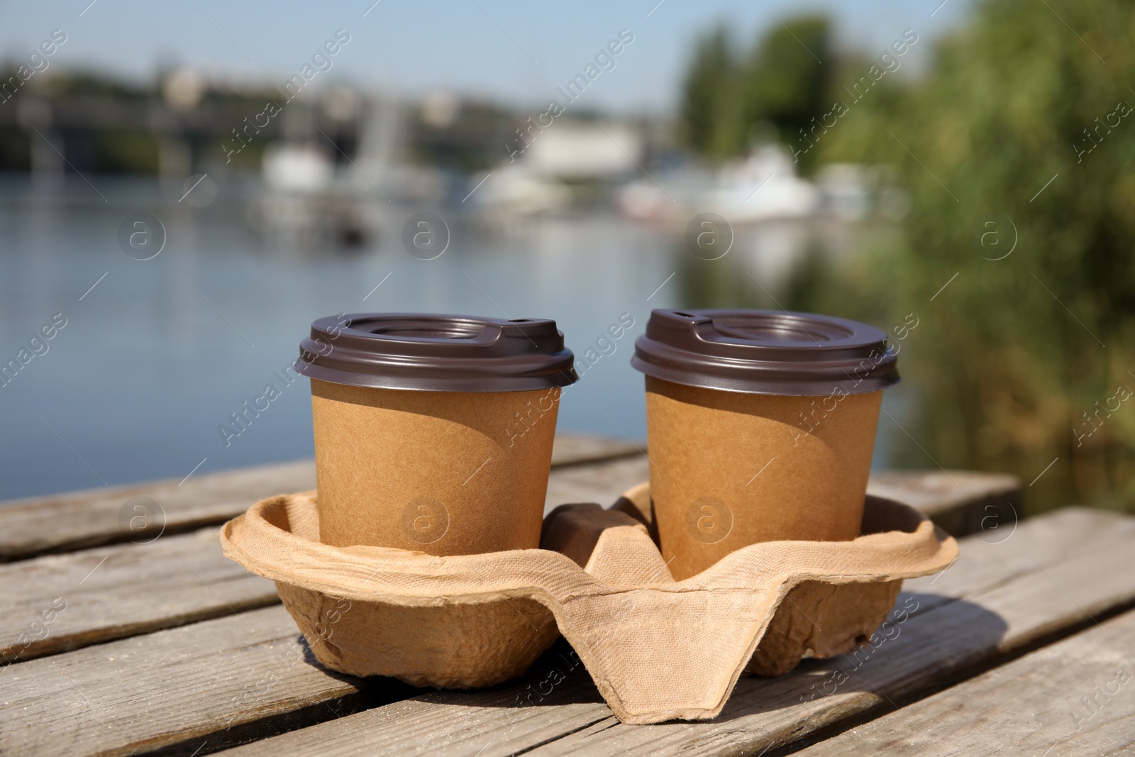 Photo of Takeaway paper coffee cups with plastic lids in cardboard holder on wooden pier near river