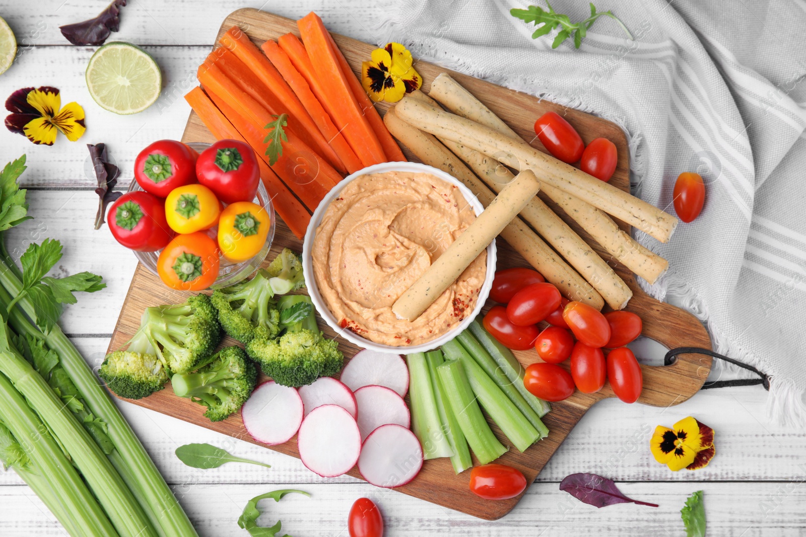 Photo of Board with delicious hummus, grissini sticks and fresh vegetables on white wooden background, flat lay