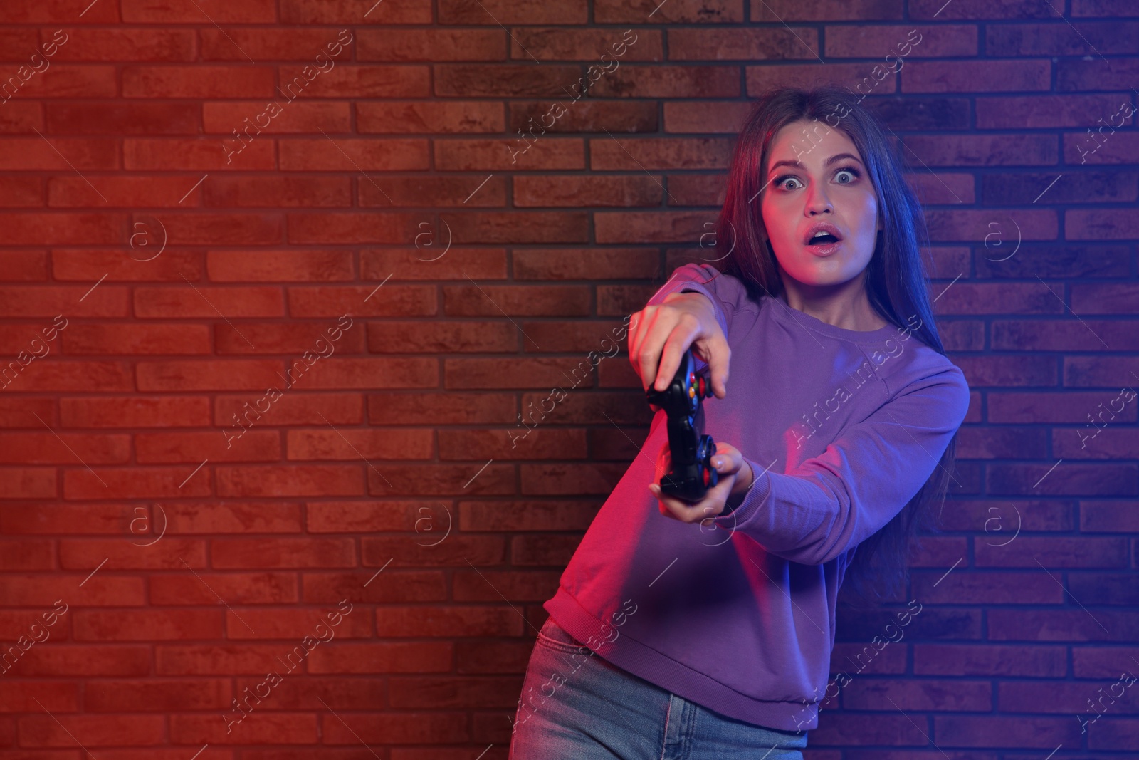 Photo of Emotional young woman playing video games with controller near brick wall. Space for text