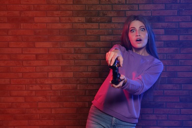 Emotional young woman playing video games with controller near brick wall. Space for text