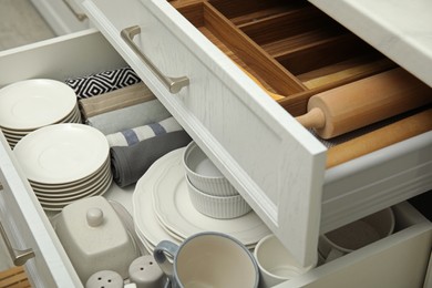 Open drawers of kitchen cabinet with different dishware, utensils and towels