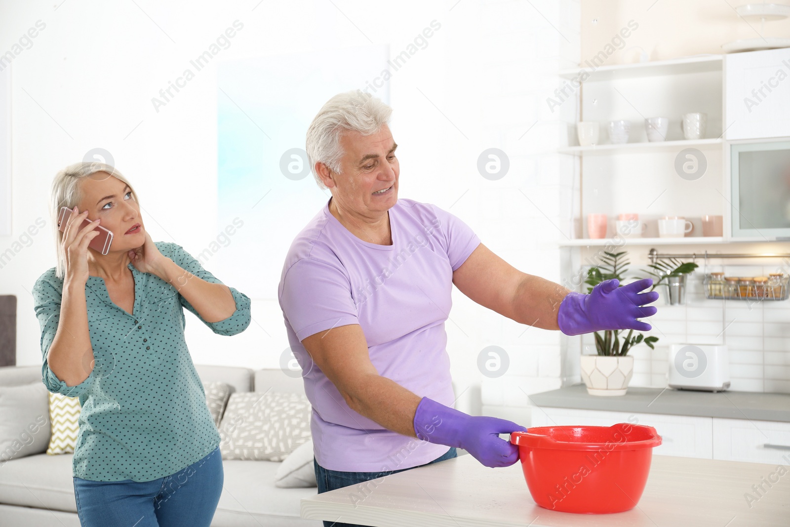 Photo of Worried mature man collecting water leaking from ceiling while his wife calling plumber at home