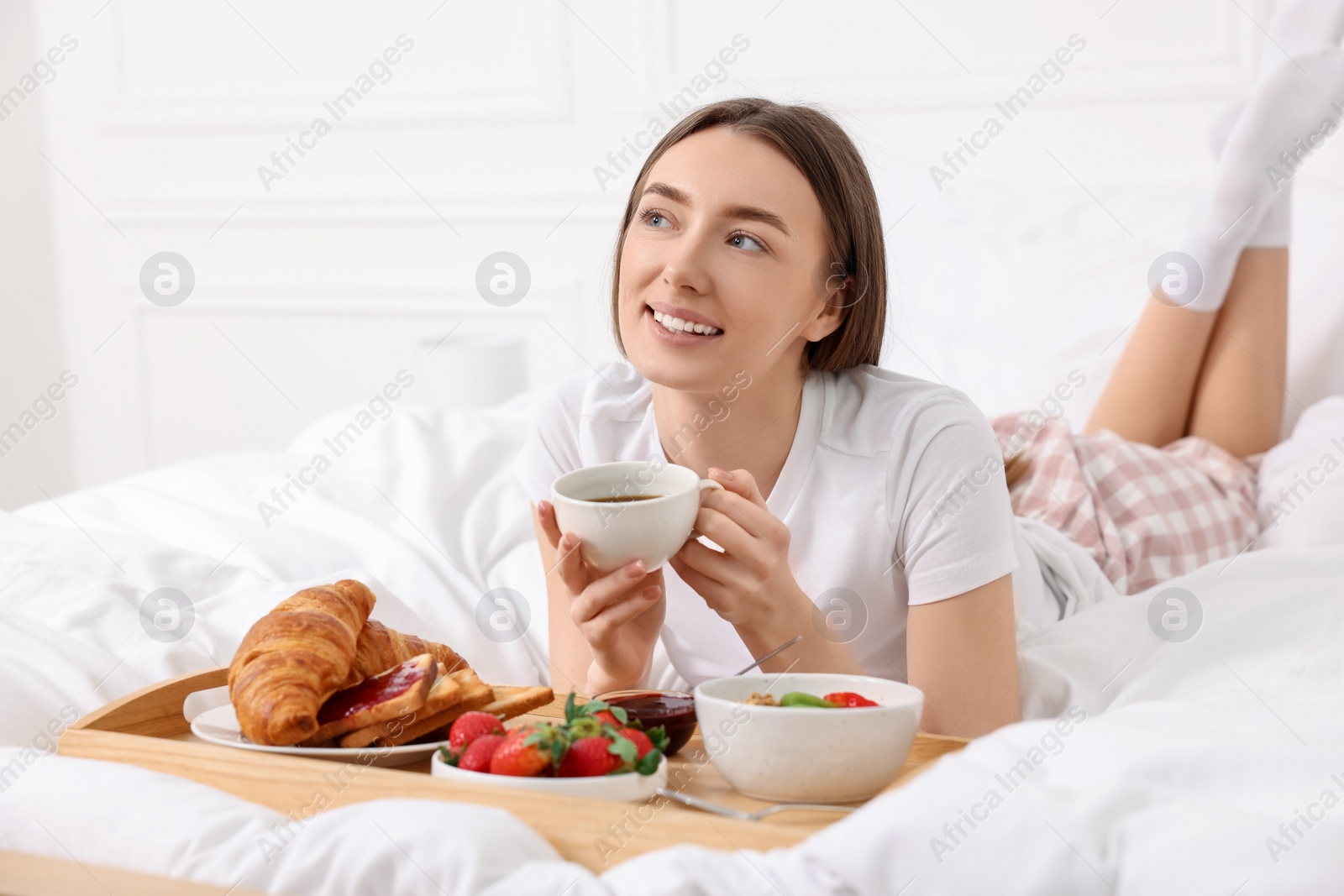 Photo of Smiling woman drinking coffee near tray with breakfast on bed