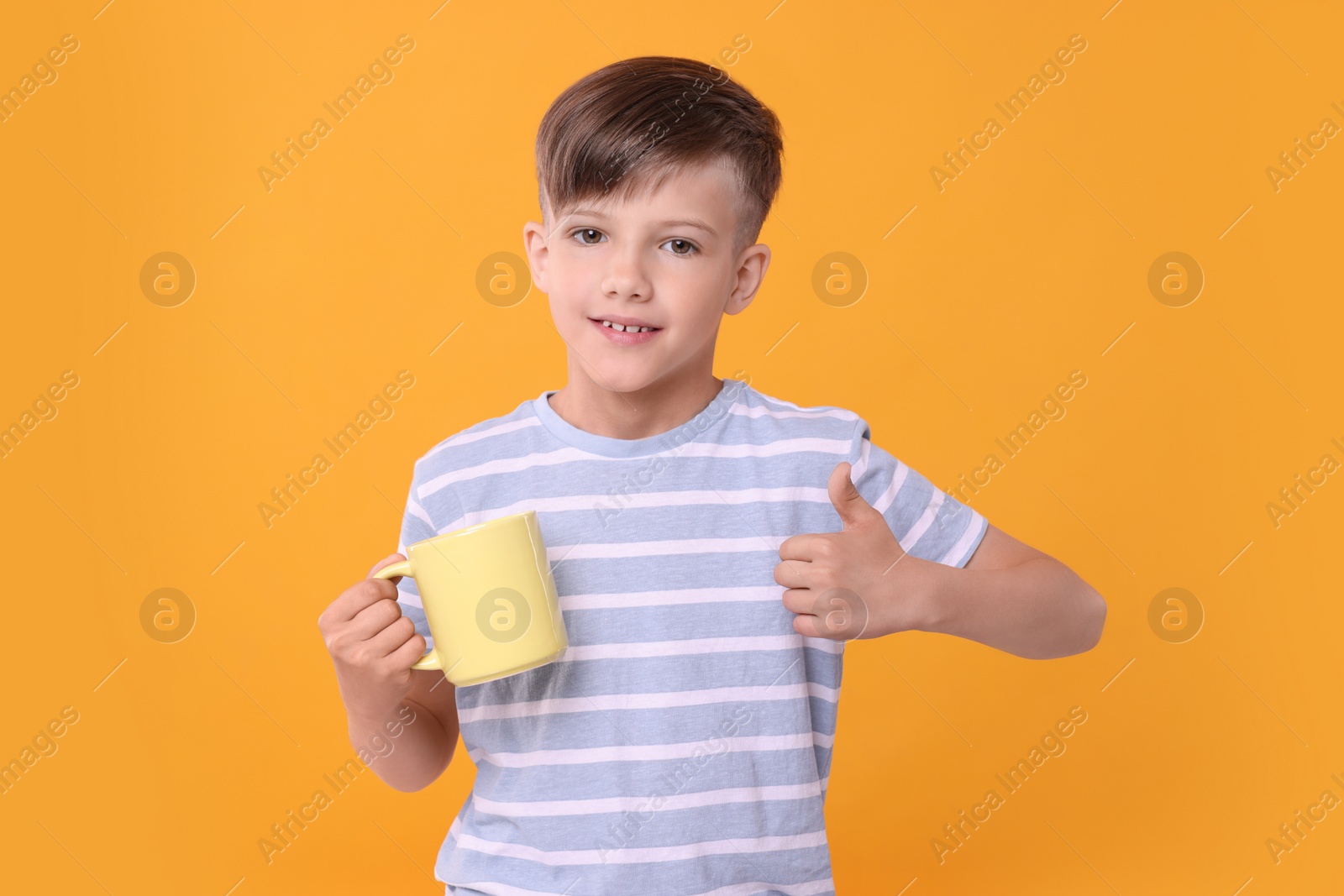 Photo of Cute boy with yellow ceramic mug showing thumbs up on orange background