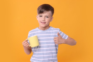 Cute boy with yellow ceramic mug showing thumbs up on orange background