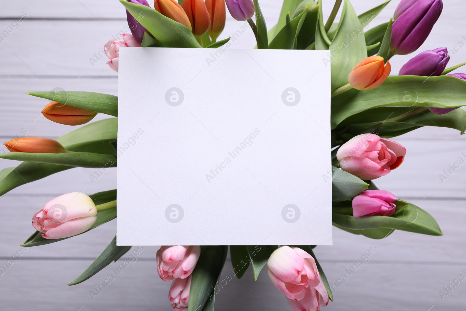 Photo of Beautiful bouquet of tulips with blank card on white wooden background, top view. Birthday celebration