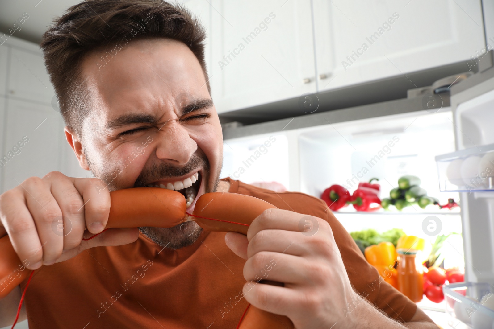 Photo of Young man eating sausages near open refrigerator, closeup