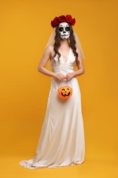 Young woman in scary bride costume with sugar skull makeup, flower crown on orange background. Halloween celebration