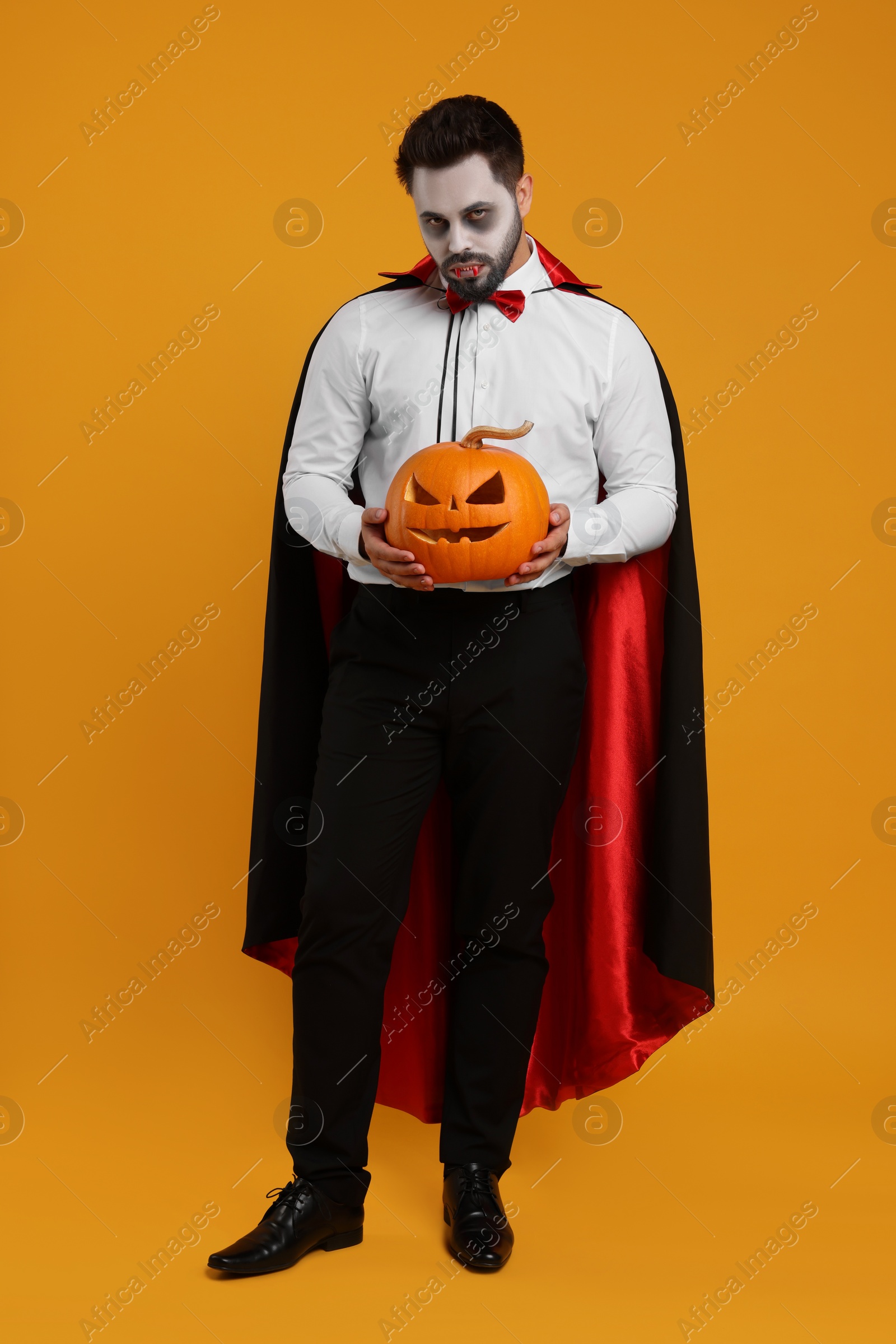 Photo of Man in scary vampire costume with fangs and carved pumpkin on orange background. Halloween celebration