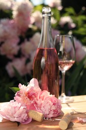 Photo of Bottle and glass of rose wine near beautiful peonies on wooden table in garden