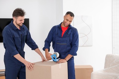 Photo of Male movers packing box with adhesive tape in house, space for text