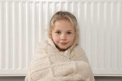 Photo of Little girl covered with knitted plaid sitting near heating radiator at home