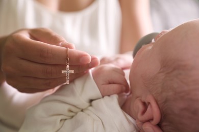 Mother holding Christian cross near newborn baby indoors, focus on hand