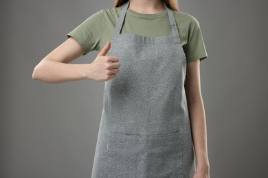 Photo of Woman in kitchen apron showing thumbs up on grey background, closeup. Mockup for design