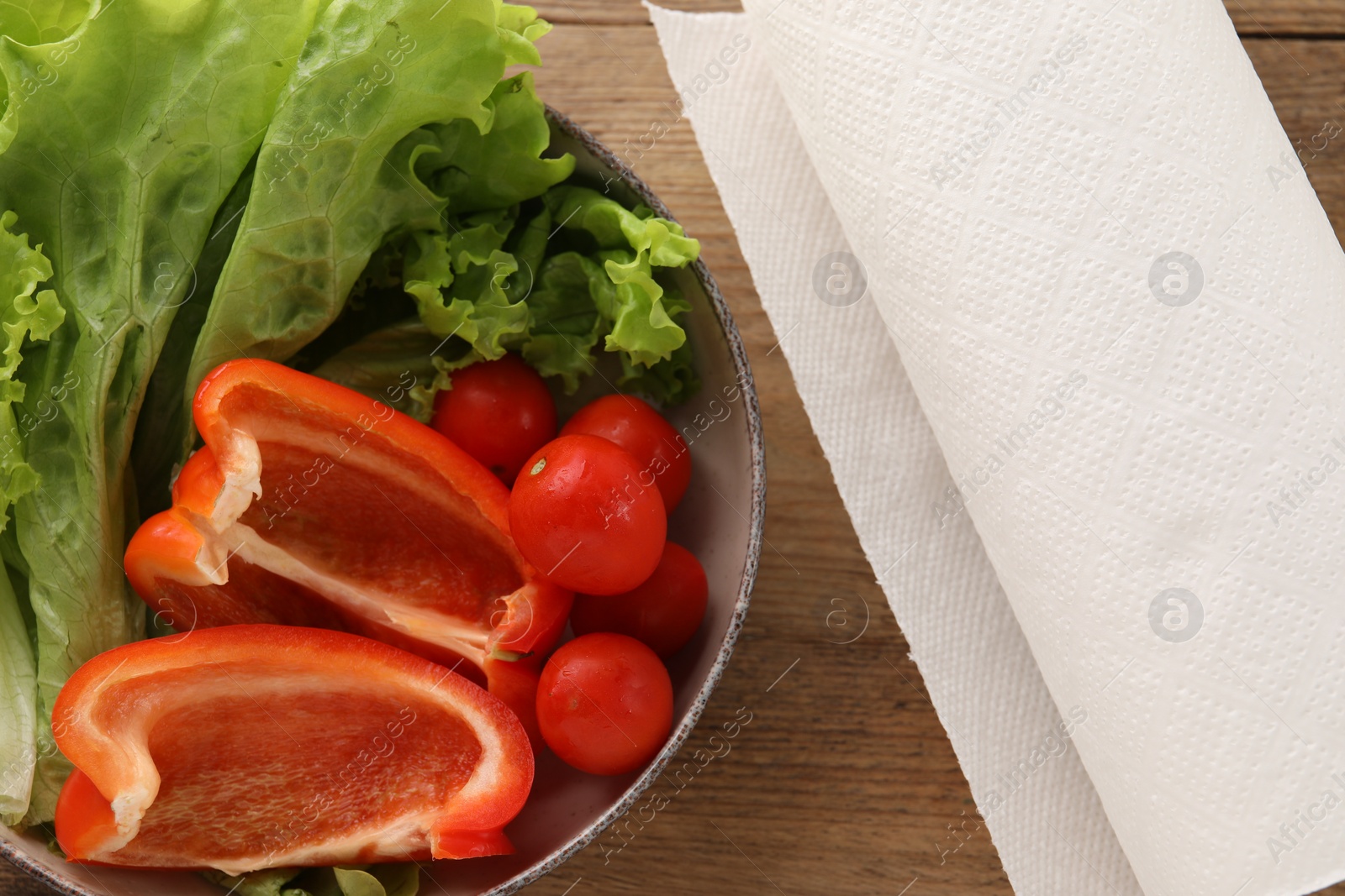 Photo of Bowl with fresh vegetables and roll of paper towels on wooden table, flat lay