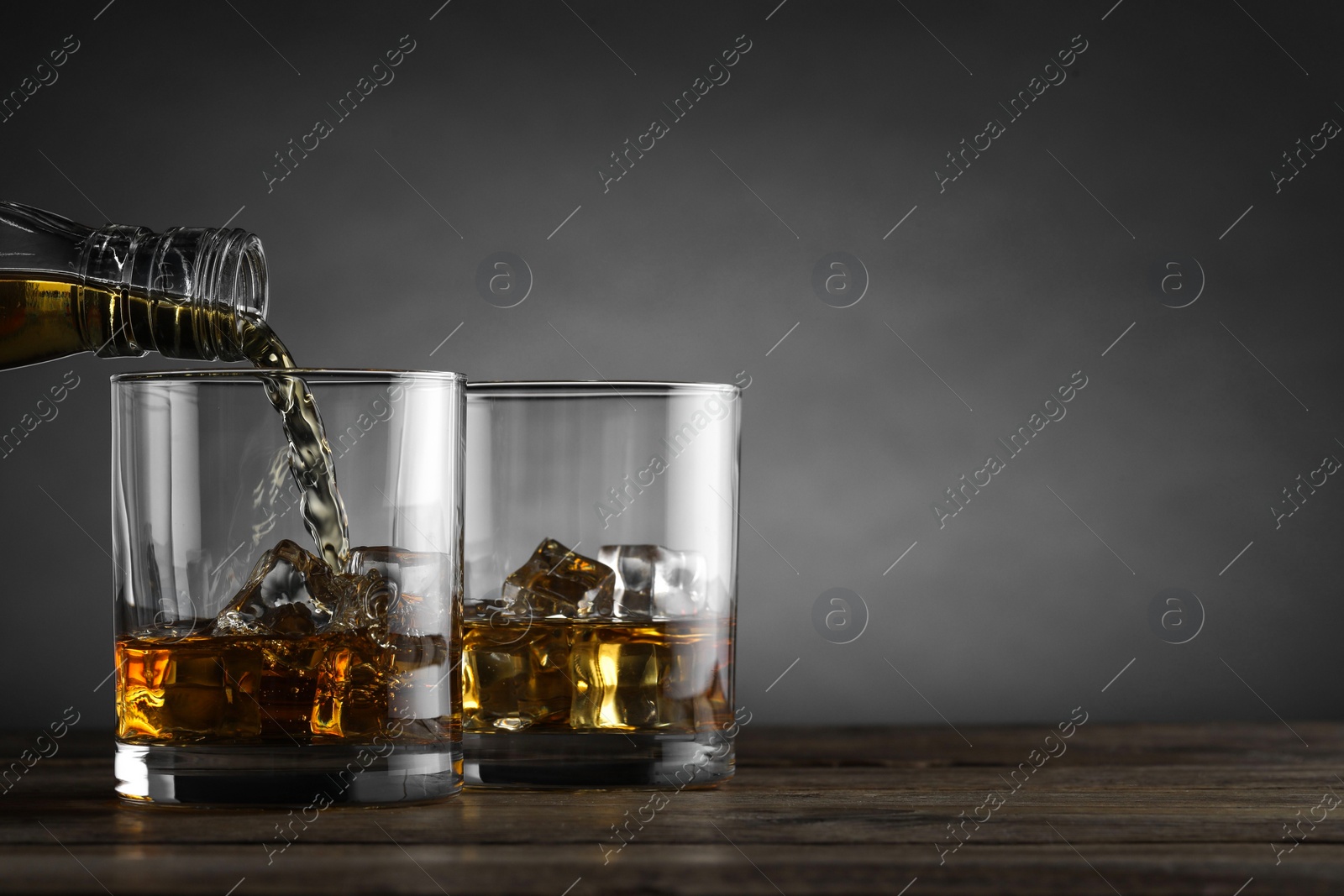 Photo of Pouring whiskey into glass with ice cubes at wooden table against grey background, space for text