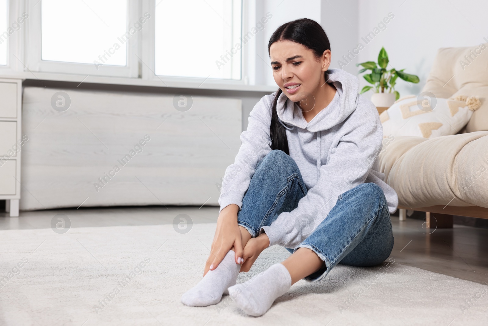 Photo of Woman suffering from ankle pain near sofa at home, space for text