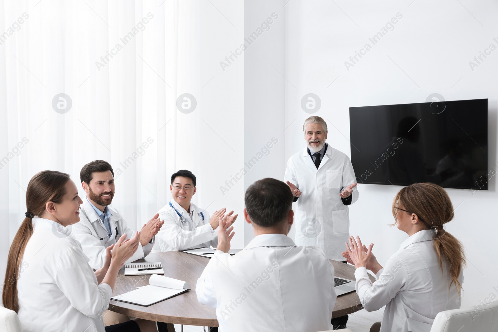 Photo of Team of doctors listening to senior speaker report near tv screen in meeting room. Medical conference