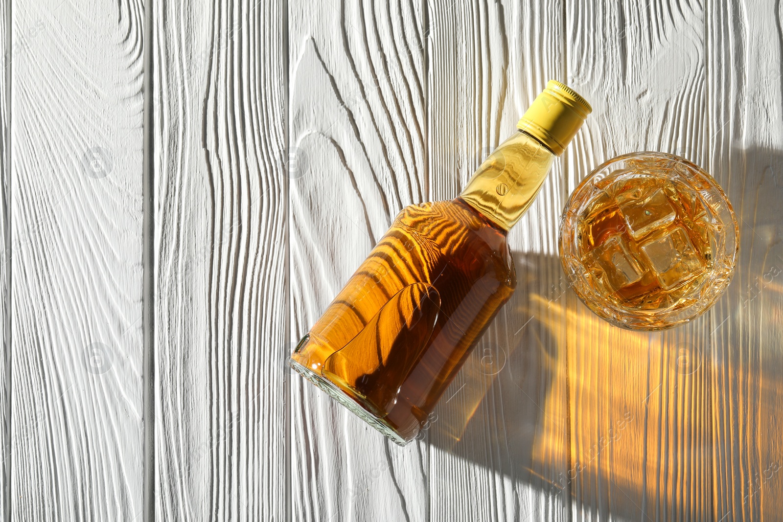 Photo of Whiskey with ice cubes in glass and bottle on white wooden table, flat lay. Space for text