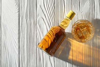 Photo of Whiskey with ice cubes in glass and bottle on white wooden table, flat lay. Space for text