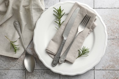 Stylish setting with cutlery, napkin, rosemary and plate on light tiled table, top view