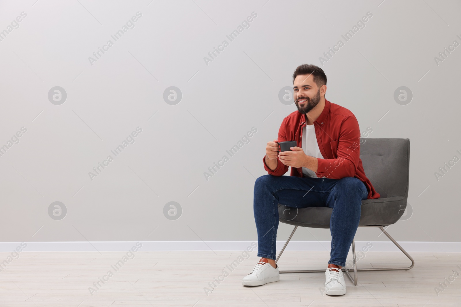 Photo of Handsome man with cup of drink sitting in armchair near light grey wall indoors, space for text