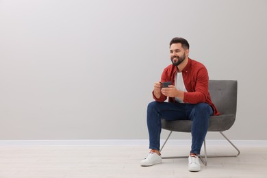 Photo of Handsome man with cup of drink sitting in armchair near light grey wall indoors, space for text