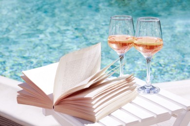 Photo of Glasses of tasty wine and open book on wooden table near swimming pool