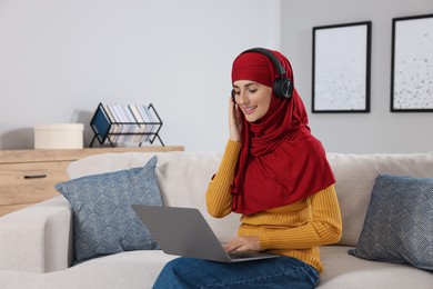 Muslim woman in headphones using laptop at couch in room