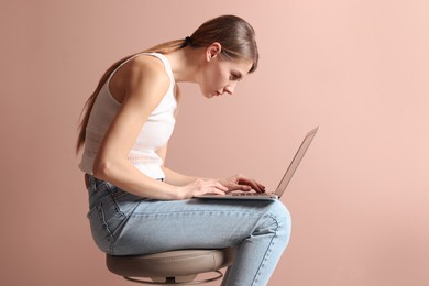 Photo of Woman with bad posture using laptop while sitting on stool against pale pink background