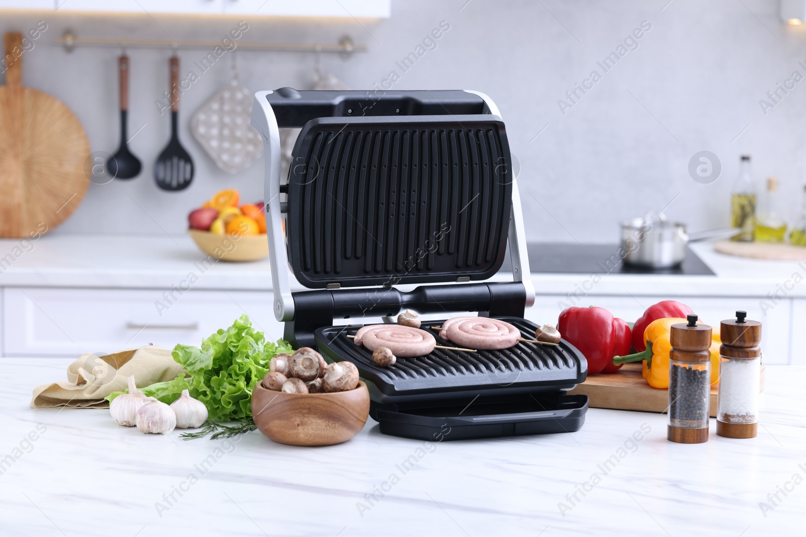 Photo of Electric grill with homemade sausages, mushrooms, vegetables and spices on white marble table in kitchen