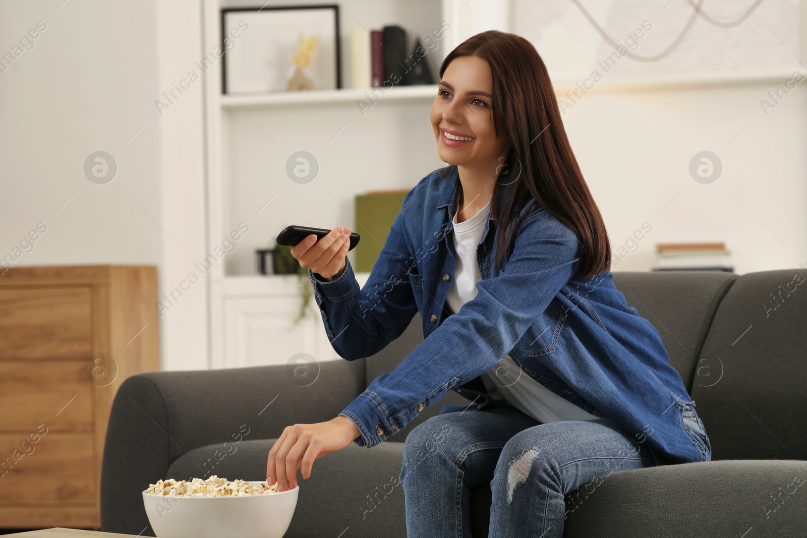 Photo of Happy woman changing TV channels with remote control on sofa at home