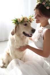 Photo of Bride and adorable Golden Retriever wearing wreath made of beautiful flowers indoors