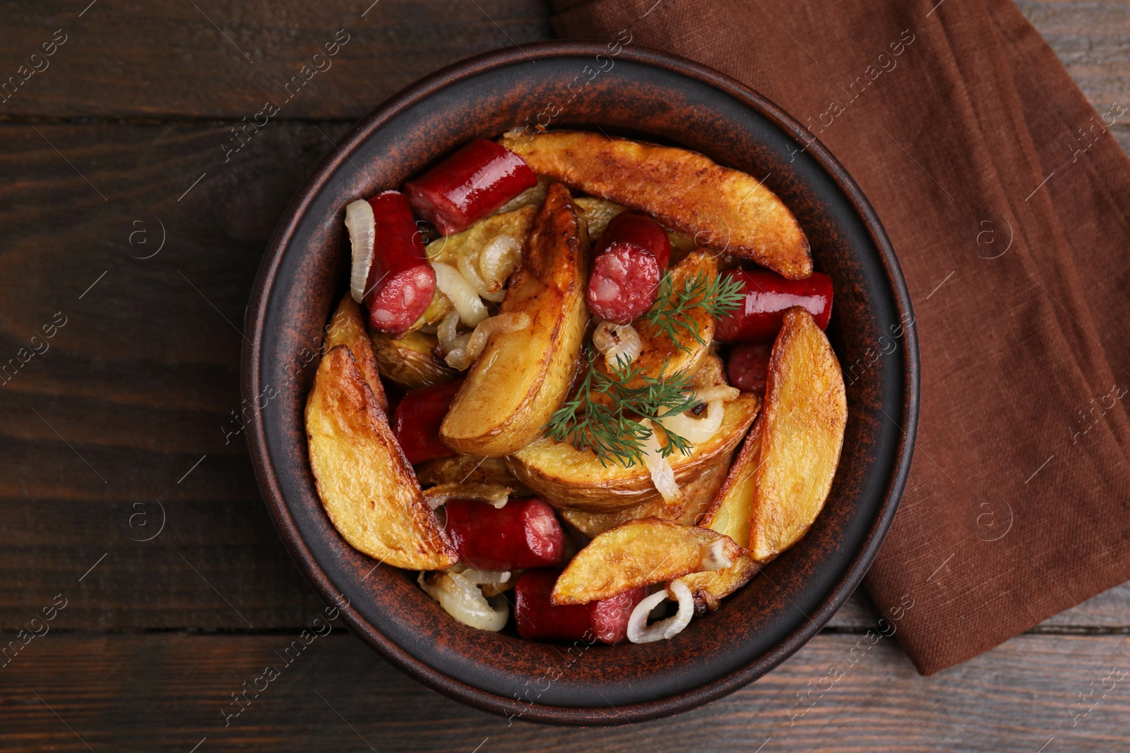 Photo of Delicious baked potato with thin dry smoked sausages, onion and dill in bowl on wooden table, top view