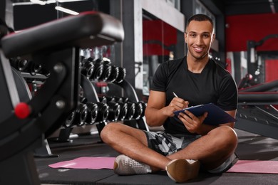 Happy trainer writing down workout plan in modern gym, space for text