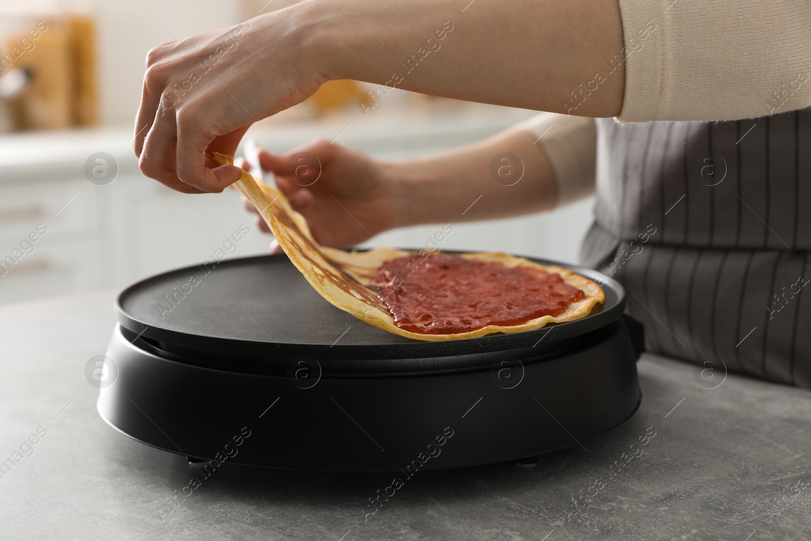 Photo of Woman cooking delicious crepe with jam on electric pancake maker in kitchen, closeup