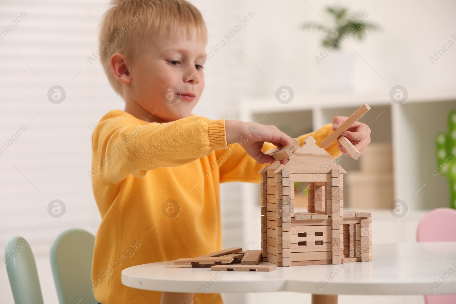 Photo of Cute little boy playing with wooden house at white table indoors. Child's toy