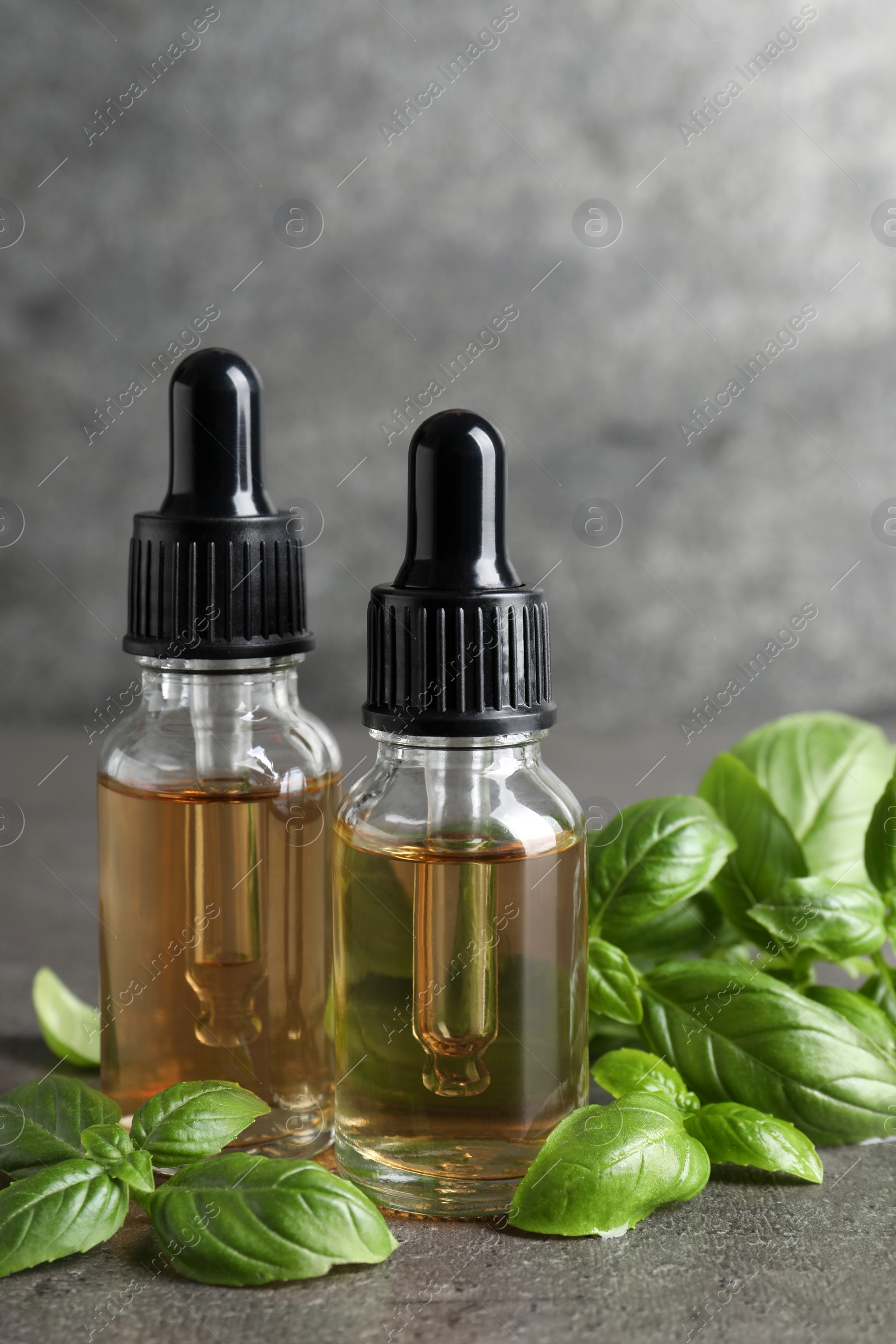 Photo of Glass bottles of basil essential oil and leaves on grey stone table