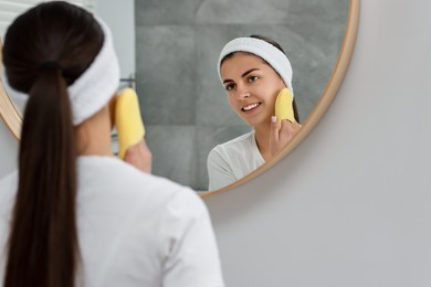 Young woman with headband washing her face using sponge near mirror in bathroom