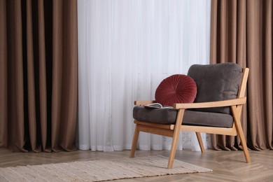 Photo of Comfortable armchair with cushion near window indoors, space for text. Interior design