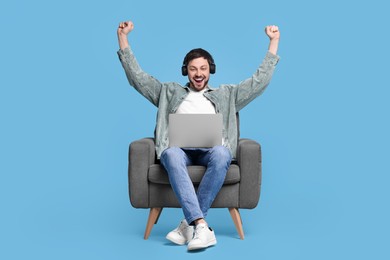 Happy man with laptop and headphones in armchair on light blue background