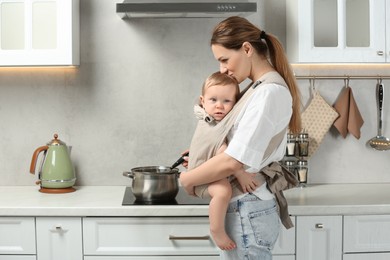 Photo of Mother holding her child in sling (baby carrier) while cooking in kitchen