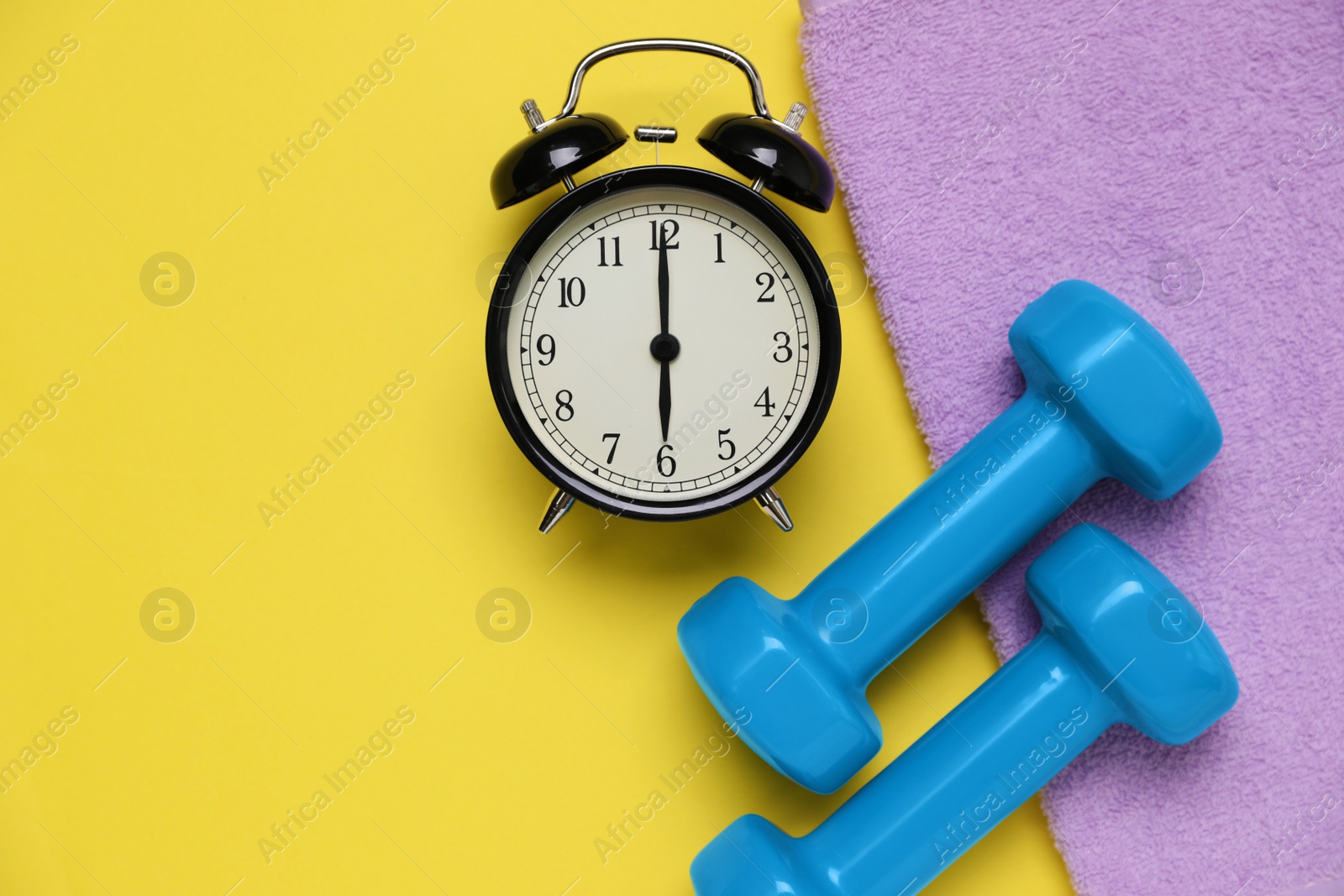 Photo of Alarm clock, towel and dumbbells on yellow background, flat lay with space for text. Morning exercise