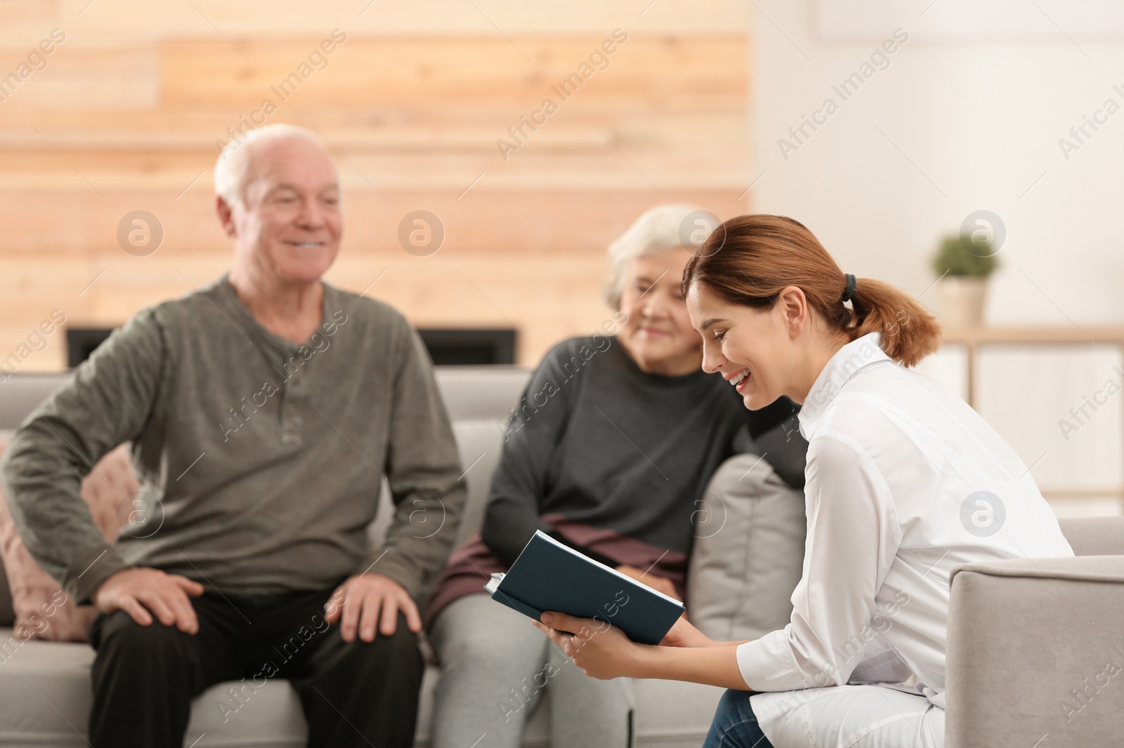Photo of Female caregiver reading book to elderly spouses in living room