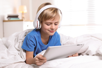 Photo of Cute little boy with headphones and tablet listening to audiobook in bed at home